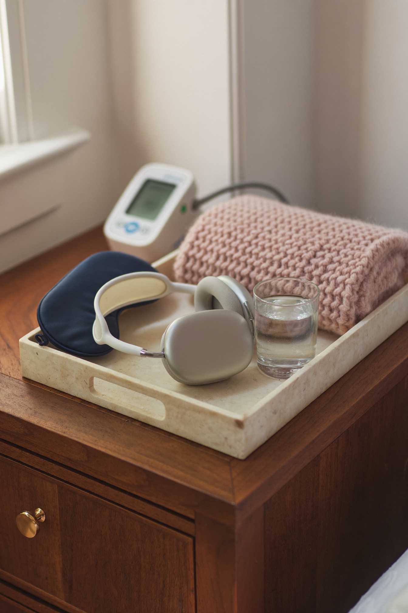 A session tray on a bedside table with headphones, an eye mask, a glass of water and a folded blanket, beside a blood pressure monitor